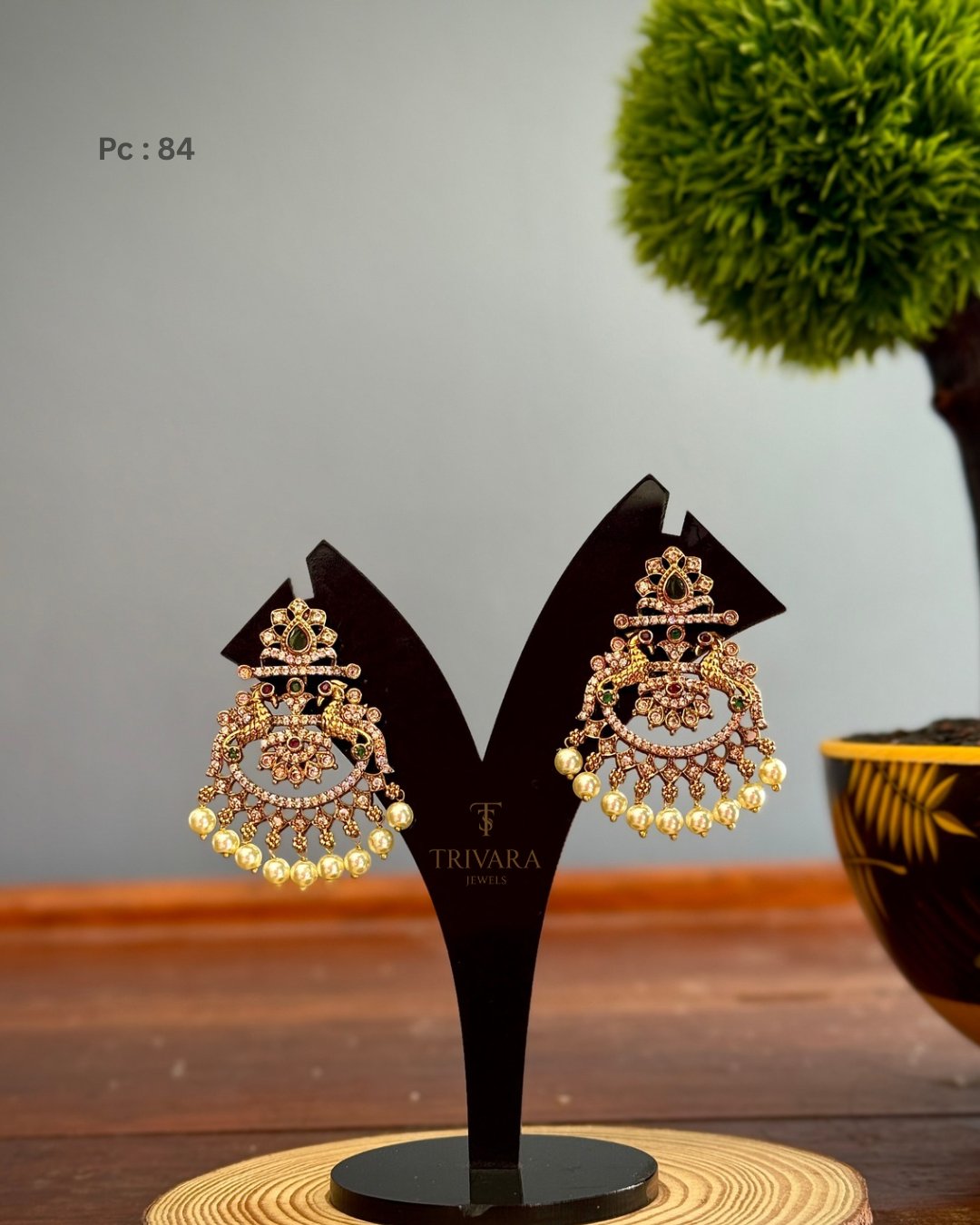 Elegant traditional Indian imitation jhumka earrings with intricate pink and green stone work, pearl drops, and antique gold finish, displayed on a Trivara Jewels stand. Perfect for weddings, festivals, and ethnic fashion styling